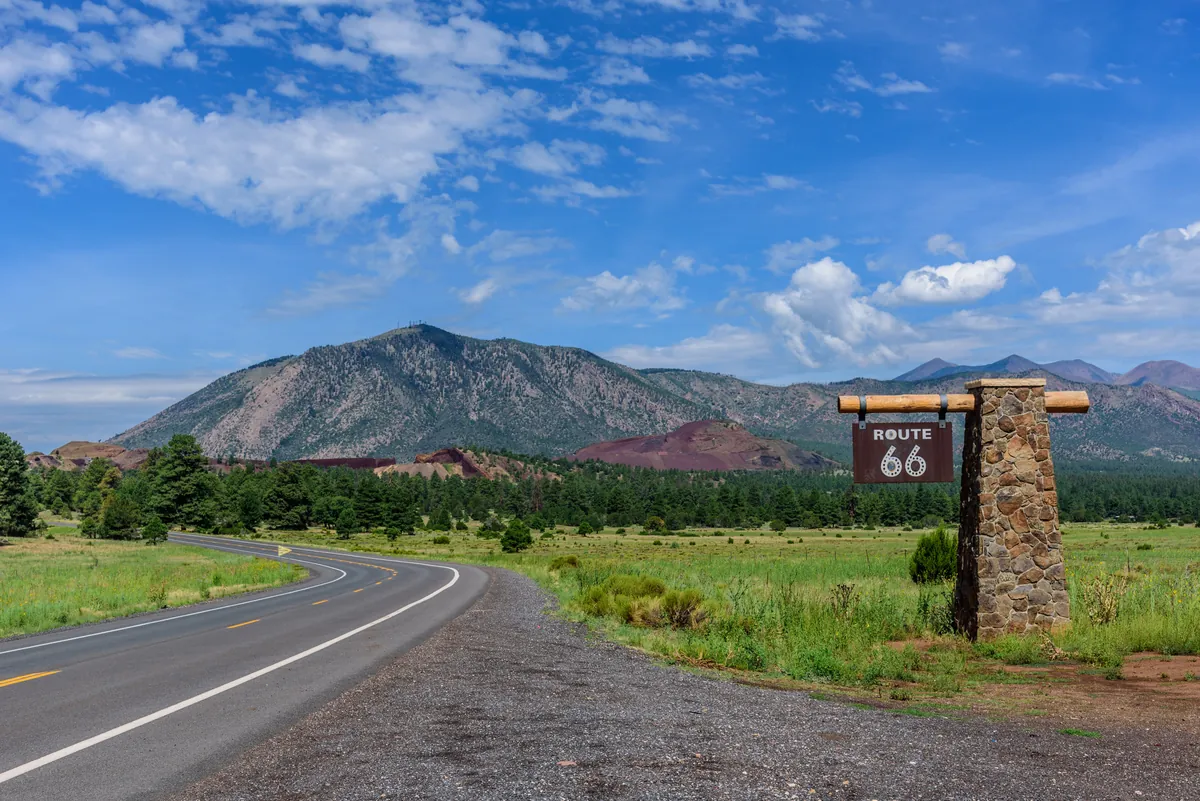 A two-lane stretch of Route 66 curving through northern Arizona, with a stone Route 66 marker and the San Francisco Peaks beyond.